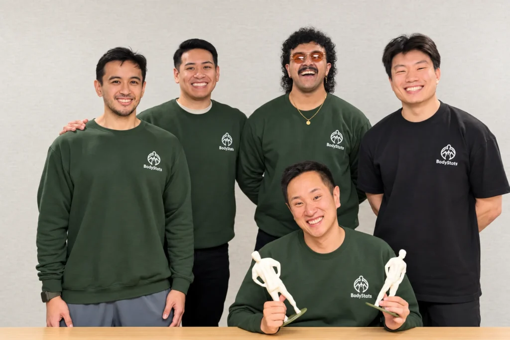 Five members of the BodyStats team in BodyStats sweatshirts and tees, one seated holding two 3D-printed body figures